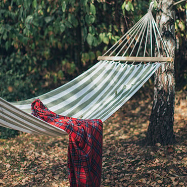 Red plaid blanket sitting on a white and green striped hammock hanging between two trees in a wooded area